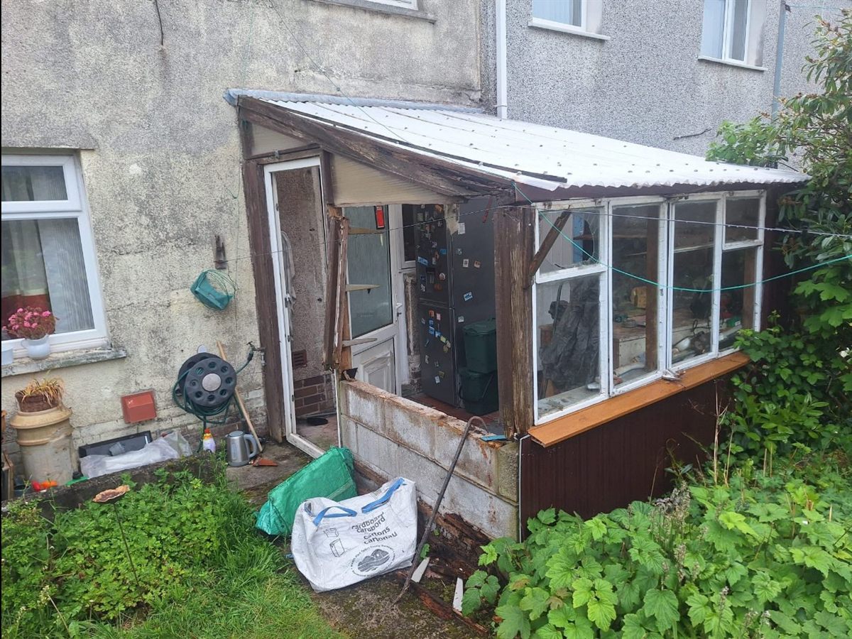 Rotten timber lean-to porch with a cracked corrugated roof, failing single glazing and a collapsing base